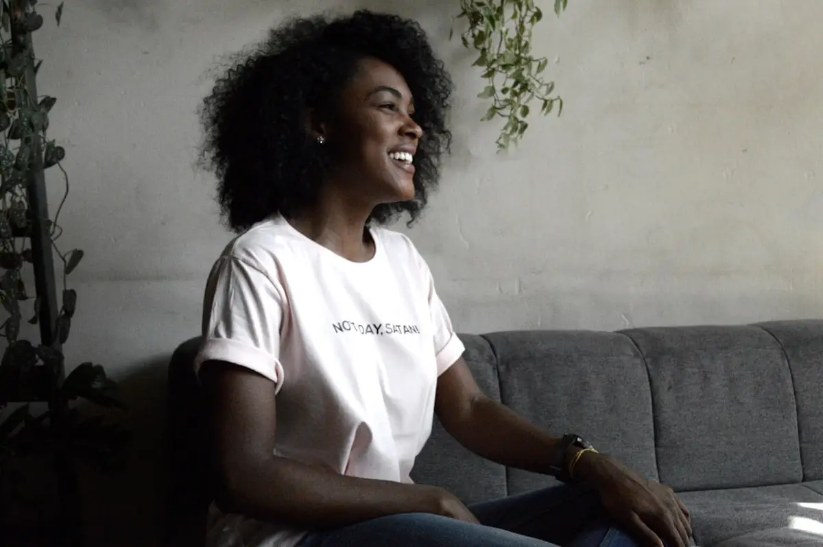 A woman caught laughing in conversation — warm soft daylight, calm sofa-side framing, a hanging plant to the side.