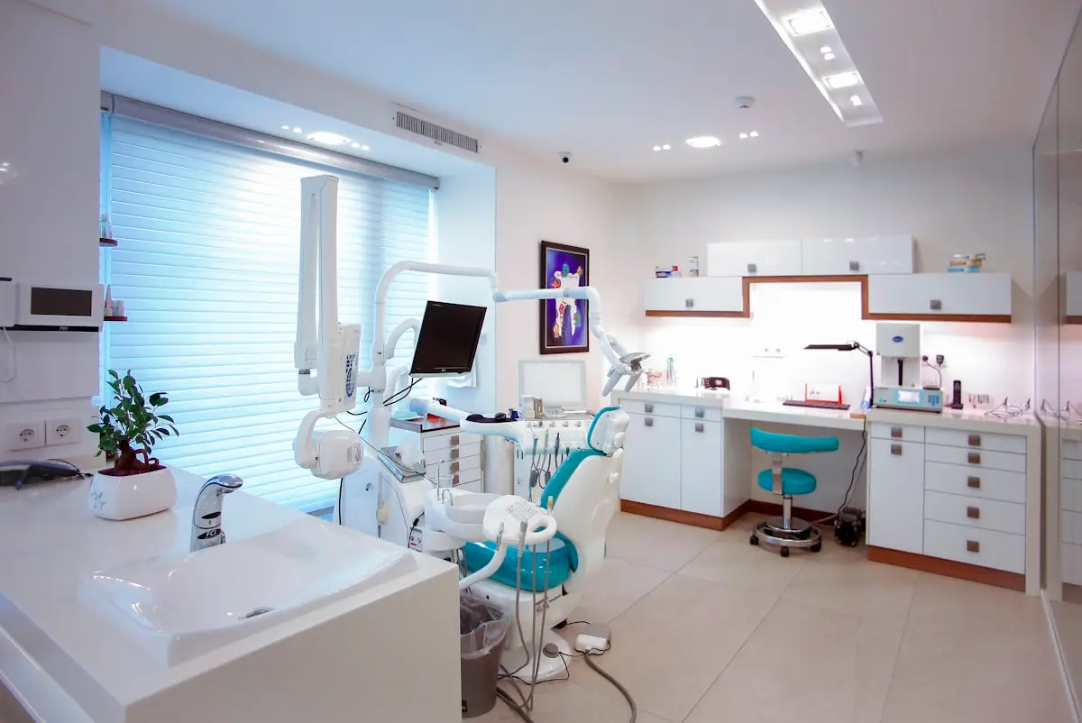 A bright, modern dental treatment room — pale teal dental chair, white wall storage, daylight through window blinds, a small plant on the counter. No patient in view, no clinical close-up. The room reads as calm and clean, not surgical.